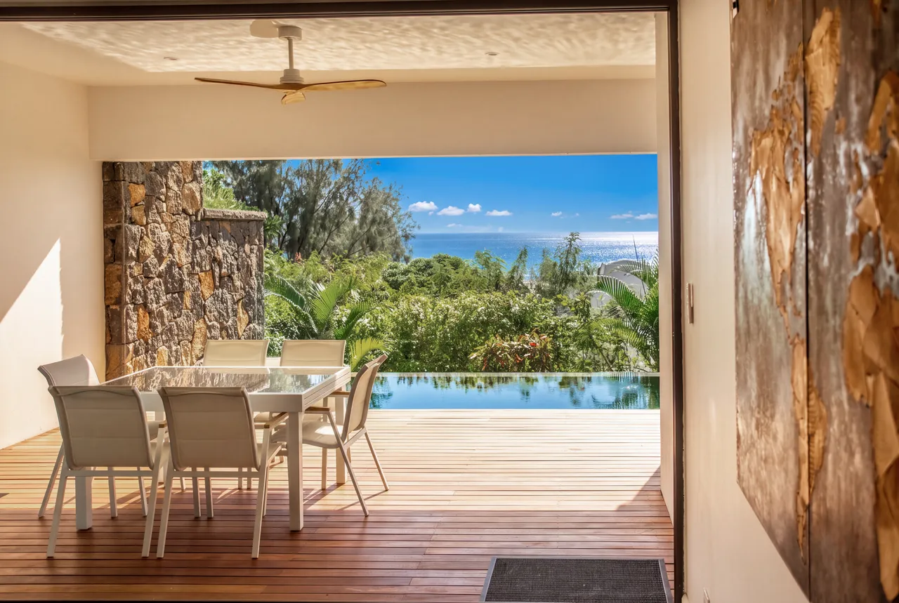 Ouverture sur une terrasse en bois avec vue sur la mer. Table et chaises pour repas en plein air.