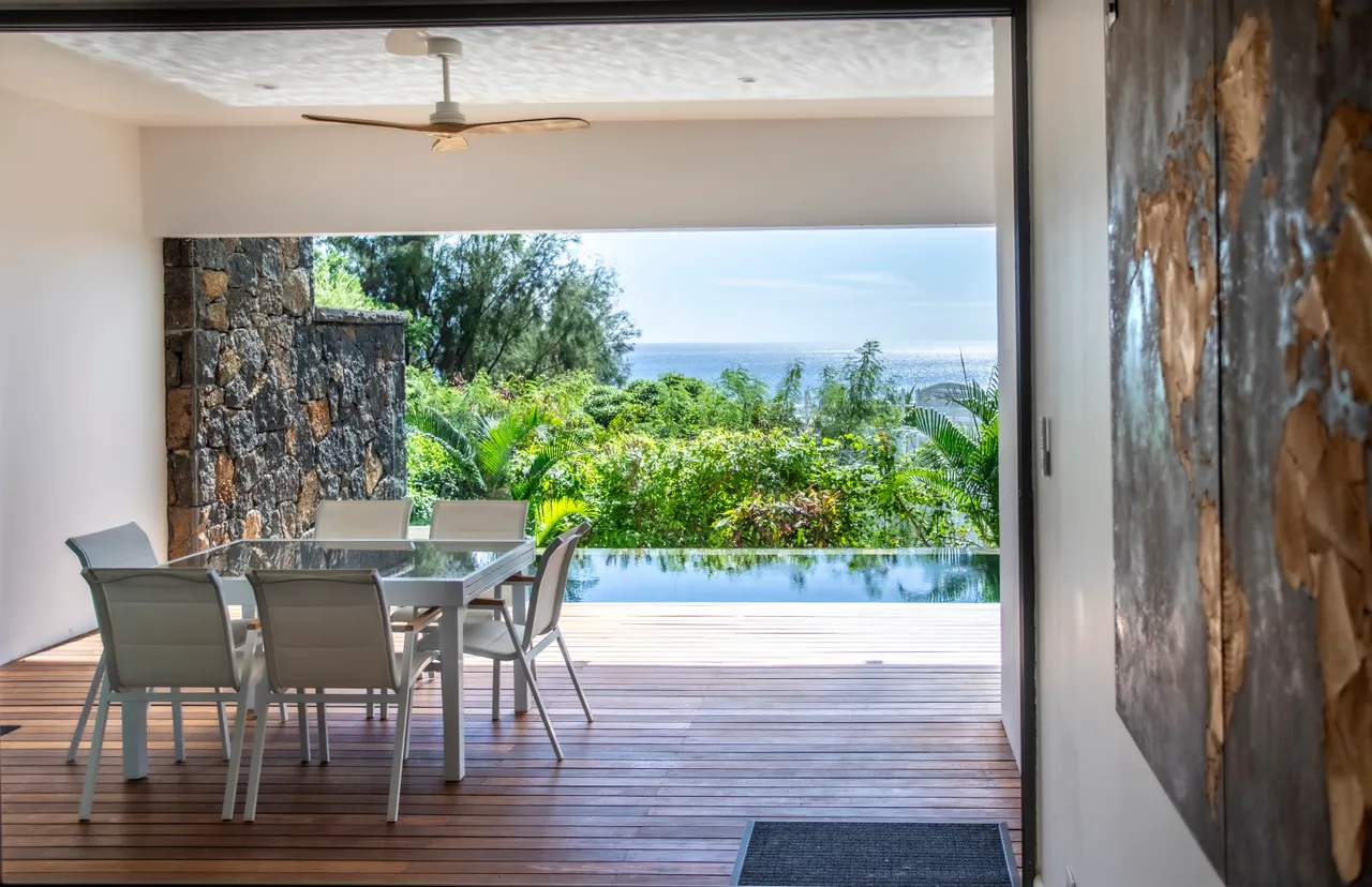 Ouverture sur une terrasse en bois avec vue sur la mer. Table et chaises pour repas en plein air.