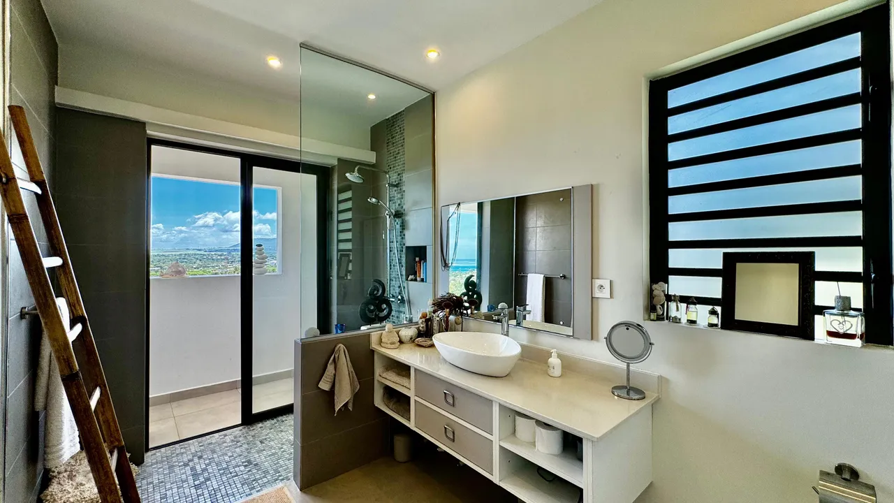 Salle de bain spacieuse avec meuble vasque et grande miroir. Fenêtres offrant une vue sur la mer.
