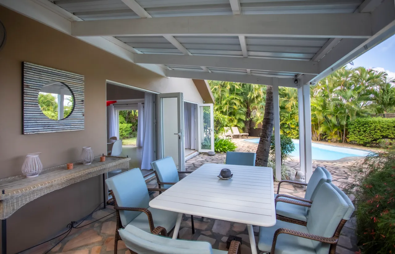 Espace de la galerie avec table et chaises pour repas en extérieur.  
Le toit ouvert offre une protection contre le soleil tout en laissant pénétrer la lumière.  
Vue sur la piscine et le jardin depuis cet espace à vivre.  
Mobilier en rotin et accessoires en métal ajoutent une touche contemporaine.  
L’aménagement permet de profiter du plein air en toute tranquillité.