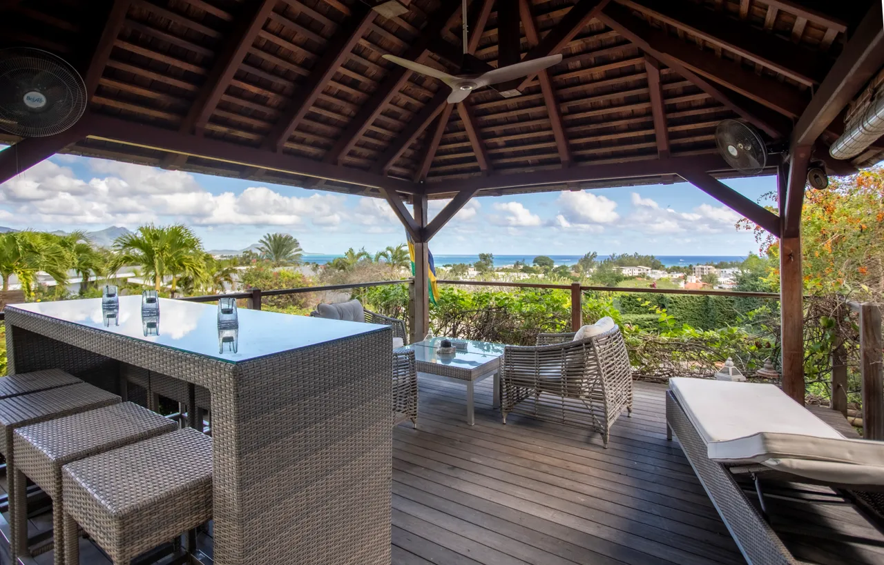 Espace de la galerie avec bar et coin détente offrant une vue sur la mer.  
Le mobilier en rotin est complété par des chaises hautes.  
Le plafond en bois ajoute une touche naturelle à l'ensemble.  
L’aménagement permet une circulation fluide entre les différentes zones.  
Le plancher en bois crée une ambiance chaleureuse et accueillante.