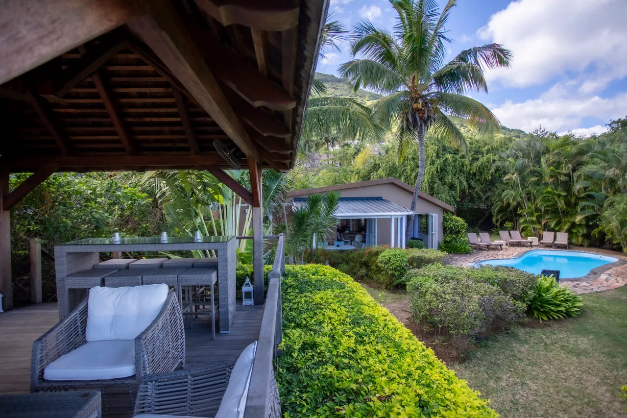 La galerie offre un espace pour se détendre avec vue sur la piscine.  
Mobilier en rotin et table à manger s'intègrent harmonieusement à l'extérieur.  
Des palmiers et des plantes tropicales entourent cet espace en plein air.  
La lumière naturelle illumine la galerie grâce à une disposition ouverte.  
L'accès direct à la piscine facilite les moments de loisirs