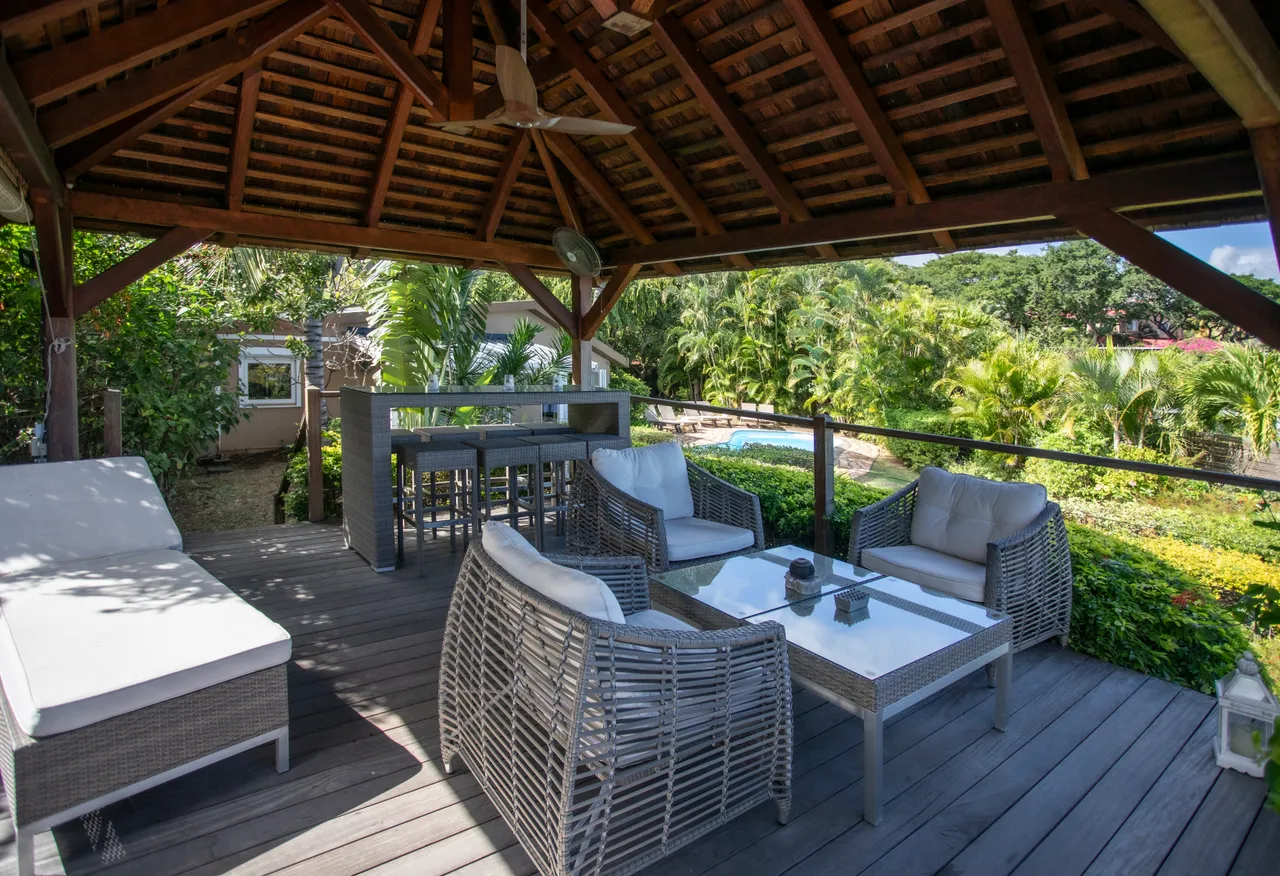 Terrasse en bois avec mobilier en rotin et table à manger.  
Le toit en bois offre une protection contre les intempéries.  
Espace extérieur entouré de verdure et de plantes tropicales.  
Coin détente avec chaises confortables idéal pour les repas.  
Vue dégagée sur le jardin depuis cet espace ombragé.