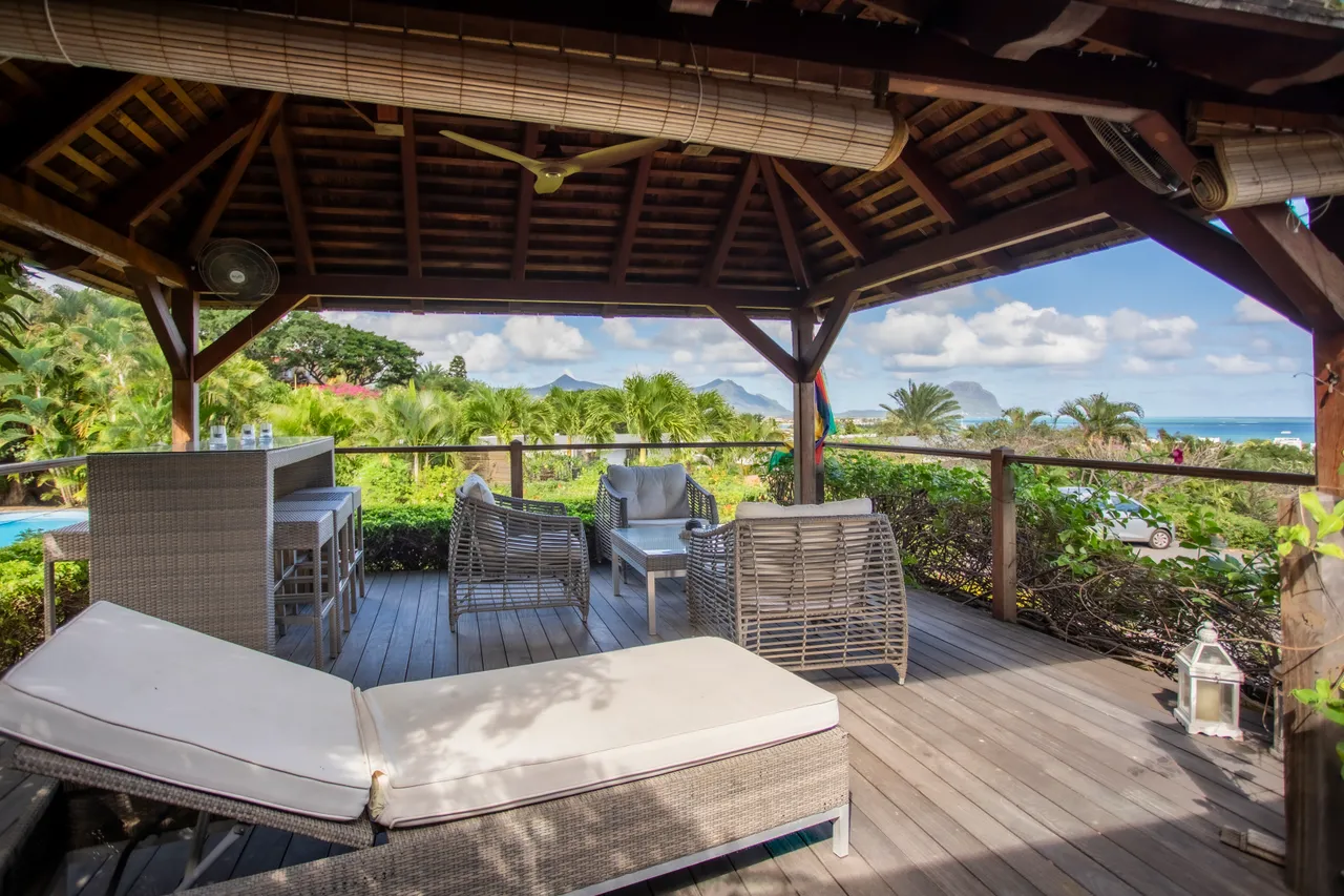 Espace de la galerie avec mobilier en rotin et chaises confortables.  
Le sol en bois apporte une touche naturelle à l'ensemble.  
Vue dégagée sur le jardin tropical et la mer environnante.  
Le toit en bois protège tout en offrant une ambiance aérée.  
Coin lounge idéal pour se détendre en extérieur.