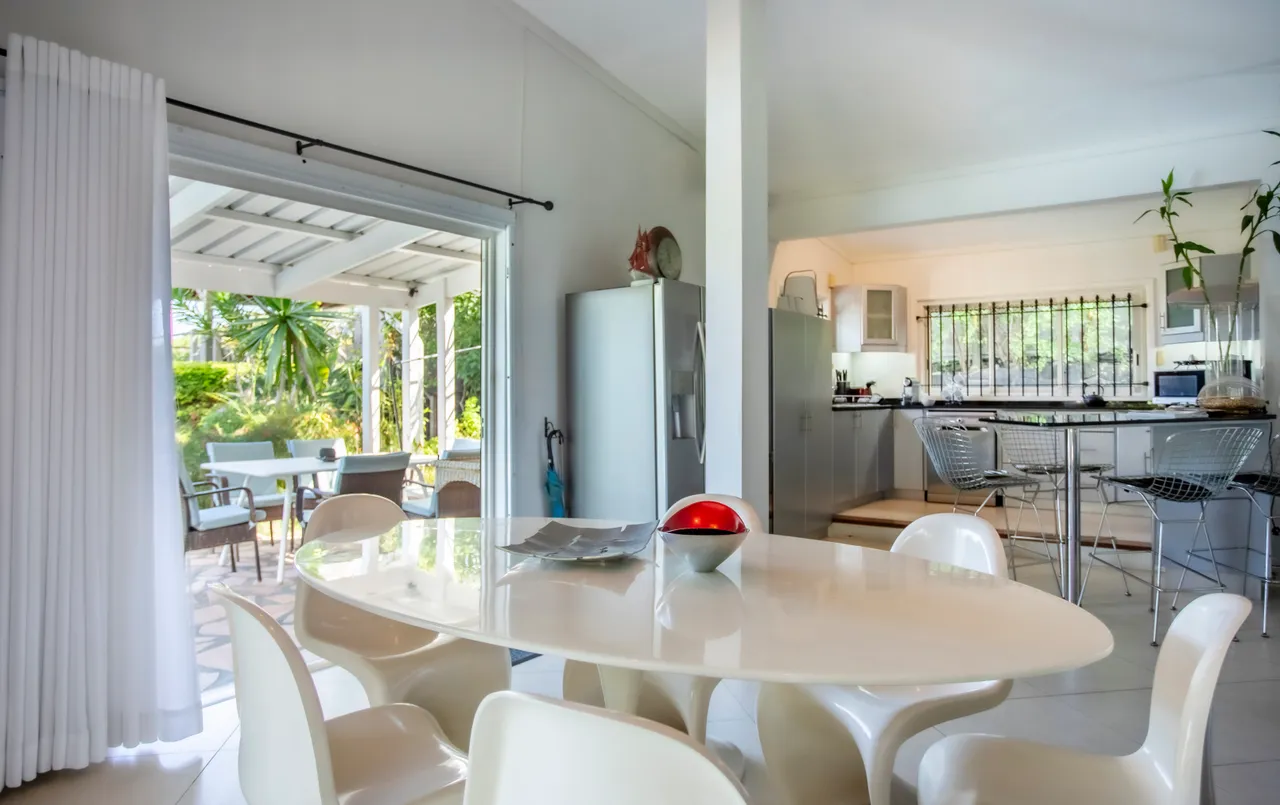 Espace de la galerie avec coin repas et grandes fenêtres donnant sur l'extérieur.  
Mobilier moderne et lumineux crée une atmosphère accueillante.  
L'agencement en open space favorise la convivialité entre les zones.  
Cuisine ouverte avec éviers et appareils électroménagers intégrés visibles.  
Végétation extérieure visible à travers les baies vitrées.