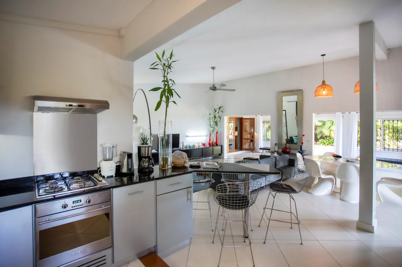 Cuisine moderne équipée de plans de travail en granit et appareils intégrés.  
Le coin repas comprend des chaises en métal autour d'une table en verre.  
Les grandes fenêtres offrent une vue sur l'extérieur, inondant l'espace de lumière.  
L'éclairage est assuré par des suspensions design au-dessus de la table.  
Le sol carrelé apporte une ambiance contemporaine à