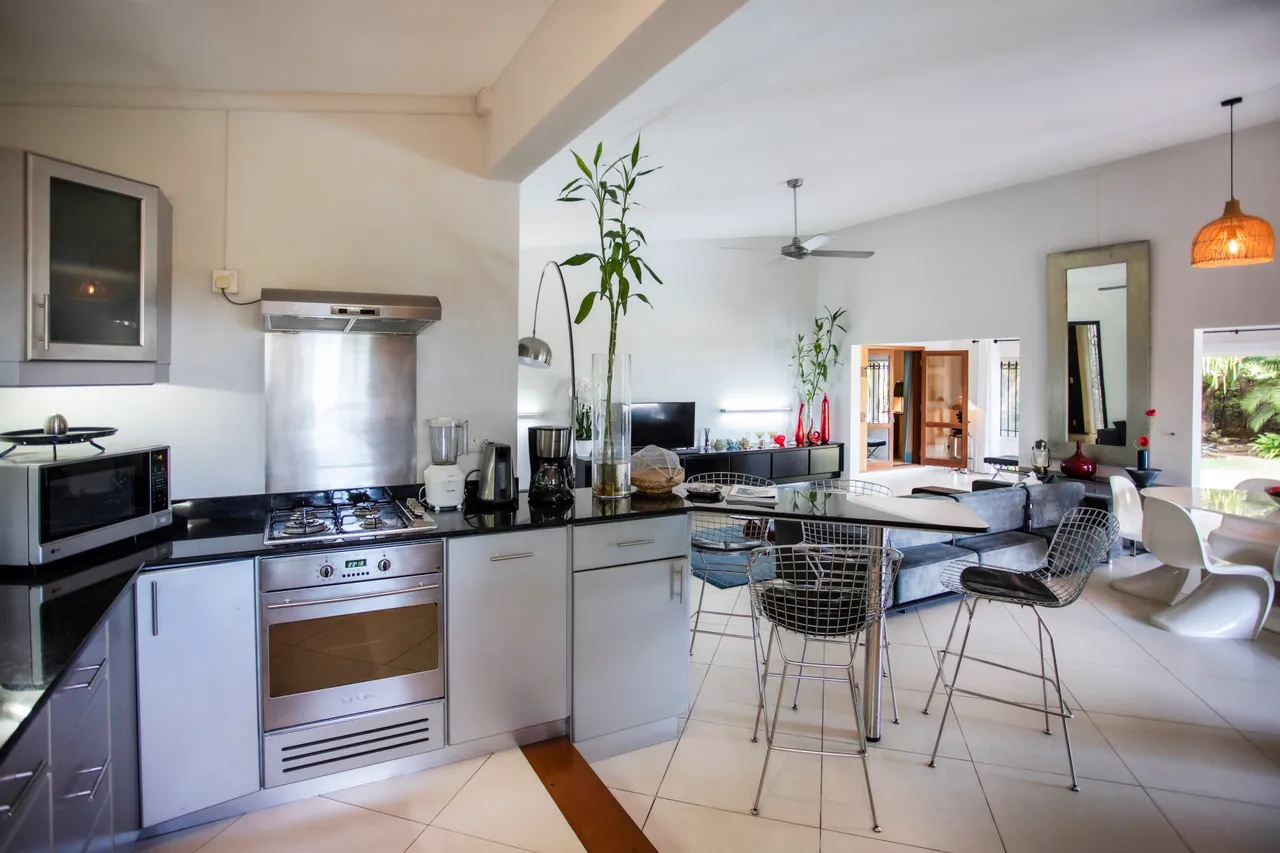 Espace de la cuisine moderne avec plans de travail en granit et appareils intégrés.  
Coin repas lumineux avec chaises en métal autour d'une table en verre.  
Les grandes fenêtres laissent entrer la lumière et offrent une vue extérieure.  
Éclairage discret grâce à des suspensions au-dessus de la table.  
Sol carrelé créant une ambiance contemporaine et aérée.