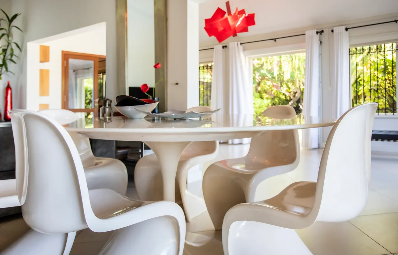 Espace de repas avec table ronde et chaises blanches au design contemporain.  
Luminaires colorés au-dessus du coin repas ajoutent une touche moderne.  
Les grandes fenêtres apportent une vue sur la végétation extérieure.  
Ambiance lumineuse créée par la lumière naturelle pénétrant dans la pièce.  
Sol carrelé contribuant à une atmosphère claire et spacieuse