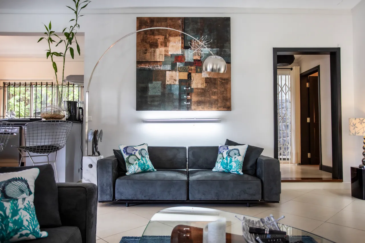 Espace de la galerie avec canapés gris et tableau moderne au mur.  
Un éclairage discret met en valeur les éléments décoratifs.  
Coin détente lumineux avec accès direct à la cuisine.  
Le sol carrelé et les meubles modernes créent une ambiance contemporaine.  
La végétation extérieure est visible à travers les grandes fenêtres.