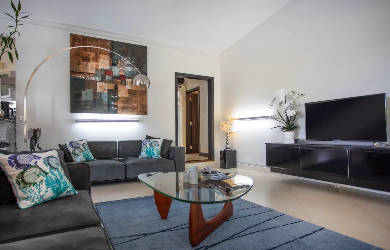 Sol carrelé clair et mobilier moderne créent une ambiance aérée dans la galerie.  
Canapés en tissu gris avec coussins colorés ajoutent du confort.  
Table basse en verre apporte une touche légère à l'ensemble.  
Luminaires contemporains assurent un éclairage fonctionnel et agréable.  
Vue dégagée sur l'extérieur grâce aux grandes fenêtres.