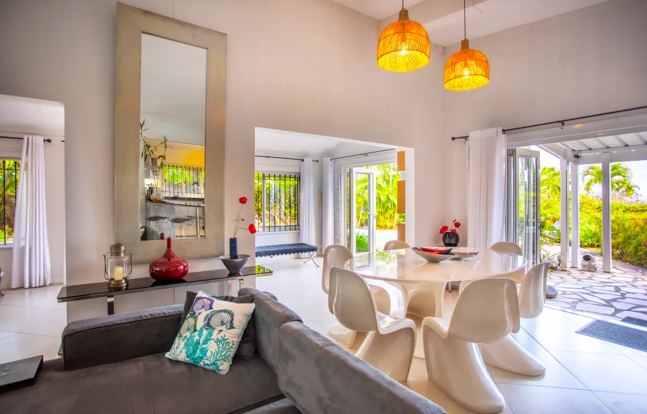 Espace de la galerie avec table à manger entourée de chaises modernes.  
Lampes suspendues en rotin ajoutent une touche chaleureuse à l'ambiance.  
Grandes baies vitrées offrent une vue sur la végétation extérieure.  
Mobilier en tissu gris et éléments décoratifs complètent l'espace.  
Le sol carrelé clair contribue à une atmosphère lumineuse
