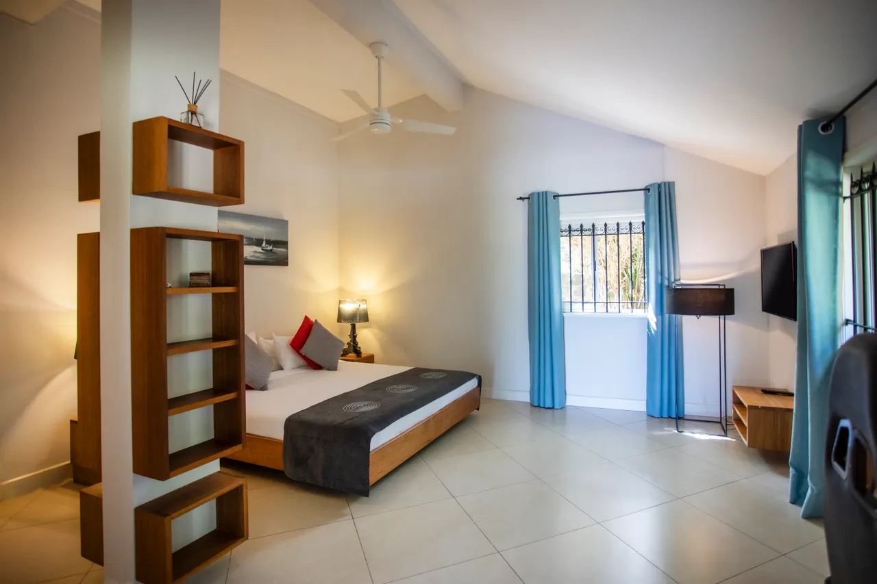 Espace chambre avec lit au design épuré et luminaires modernes.  
Les grandes fenêtres apportent une vue sur l'extérieur et la lumière naturelle.  
Étagères en bois ajoutent un espace de rangement fonctionnel.  
Ambiance calme grâce aux murs clairs et au sol carrelé.  
Coin bureau simple et pratique près de la fenêtre.