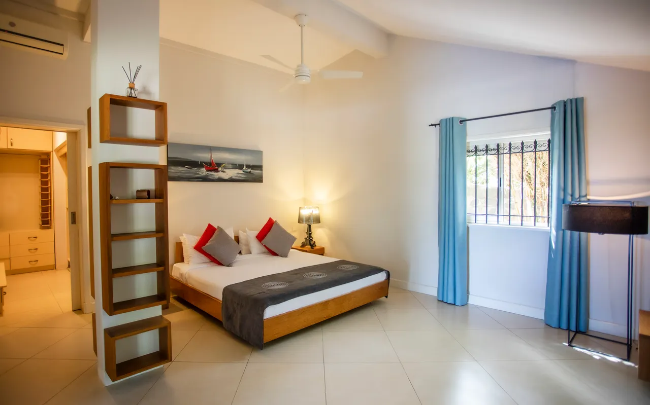 Espace chambre avec lit en bois et coussins colorés.  
Les grandes fenêtres laissent entrer la lumière et offrent une vue sur l'extérieur.  
Étagères en bois apportent un espace de rangement supplémentaire.  
Luminaires modernes créent une ambiance douce et accueillante.  
Le sol carrelé clair contribue à une atmosphère lumineuse et apaisante.
