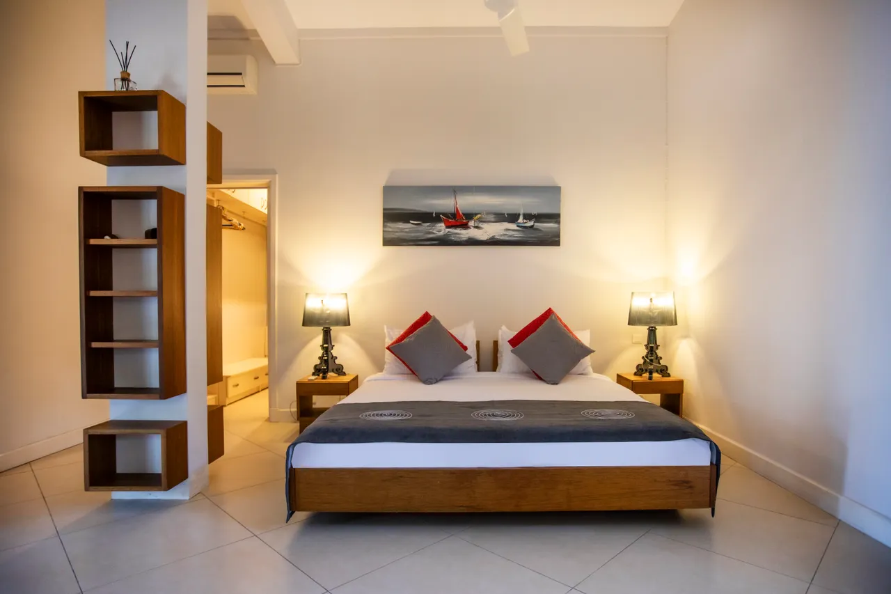 Chambre avec un lit en bois et lampes de chevet.  
Le sol en carrelage clair reflète la lumière naturelle.  
Rangement disponible dans un placard intégré à mur.  
Tableau en toile ajoutant une touche décorative à l'espace.  
Les coussins décoratifs apportent une note de couleur.  
Deux lampes de chevet fournissent un éclairage