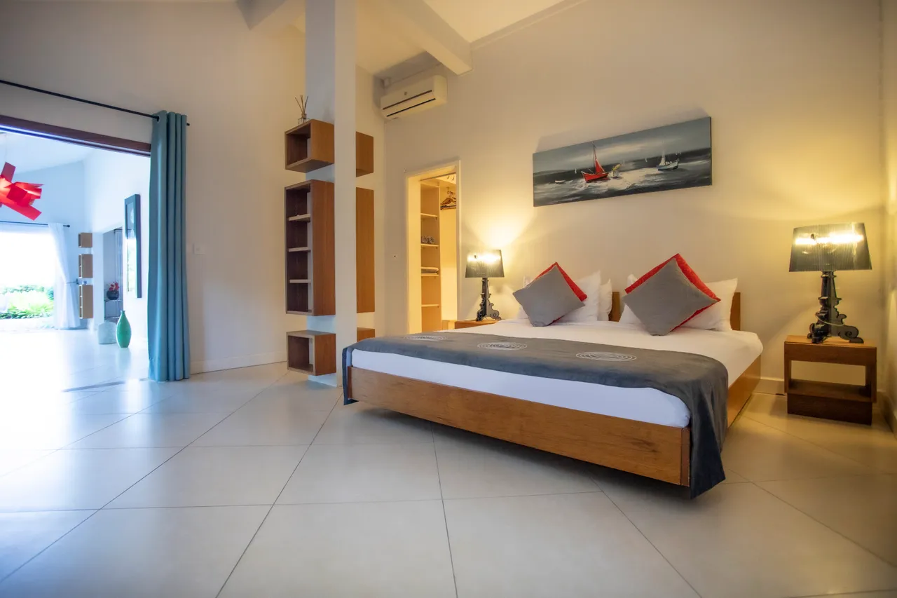 Chambre avec lit en bois et couette grise, ambiance lumineuse.  
Éclairage doux grâce aux lampes de chevet modernes.  
Grandes fenêtres offrant une vue sur l'extérieur et lumière naturelle.  
Étagères en bois ajoutent un espace de rangement pratique.  
Sol carrelé clair contribuant à une atmosphère spacieuse.