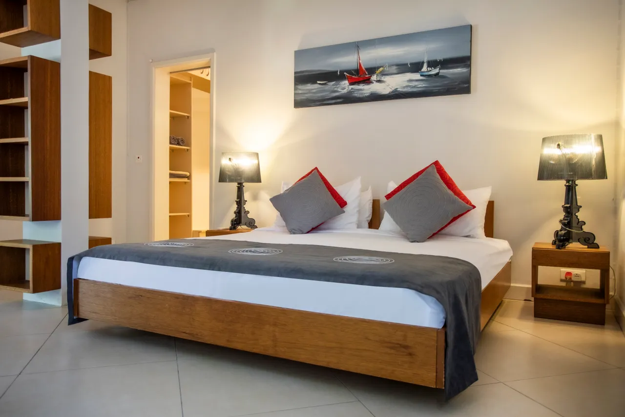Chambre avec lit en bois et décor minimaliste, ambiance claire.  
Luminaires modernes de chaque côté du lit ajoutent une touche fonctionnelle.  
Les grandes fenêtres offrent une vue dégagée et une belle lumière.  
Étagères intégrées fournissent un espace de rangement pratique.  
Sol carrelé clair contribue à l'atmosphère spacieuse de la pièce.