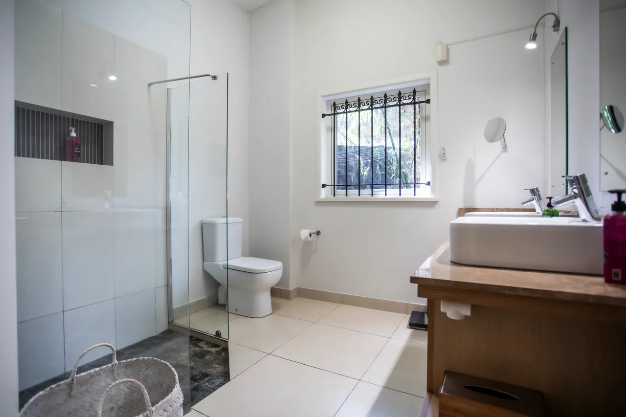 Espace salle de bain avec double vasque en bois et miroir éclairé.  
Douches à l'italienne en verre transparent créent une atmosphère moderne.  
Fenêtre avec grille offre une vue sur l'extérieur et permet l'aération.  
Sol carrelé clair et murs en faïence blanche renforcent la luminosité.  
Panier tressé apporte une touche décor