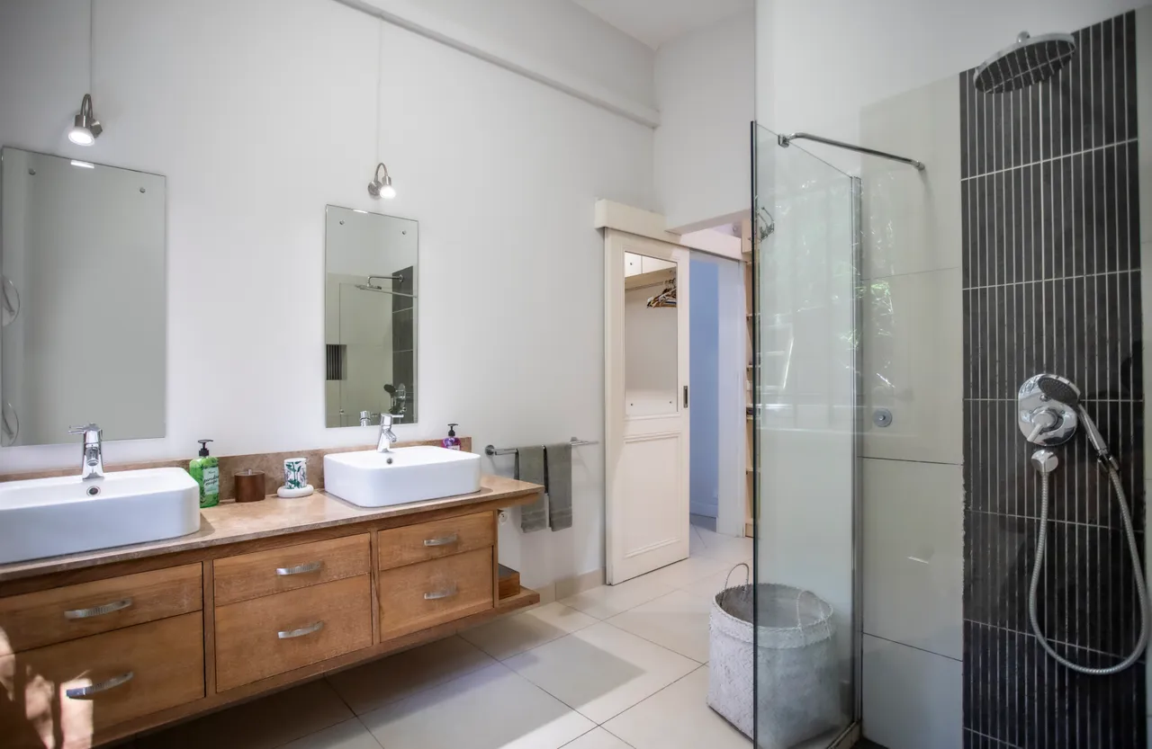 Salle de bain avec double vasque en bois et miroir éclairé au-dessus.  
Douche à l'italienne en verre offre un espace moderne et fonctionnel.  
Fenêtre avec grille permet d'aérer tout en apportant de la lumière.  
Étagères intégrées fournissent un rangement pratique pour les accessoires.  
Sol carrelé clair et murs en faïence blanche favor