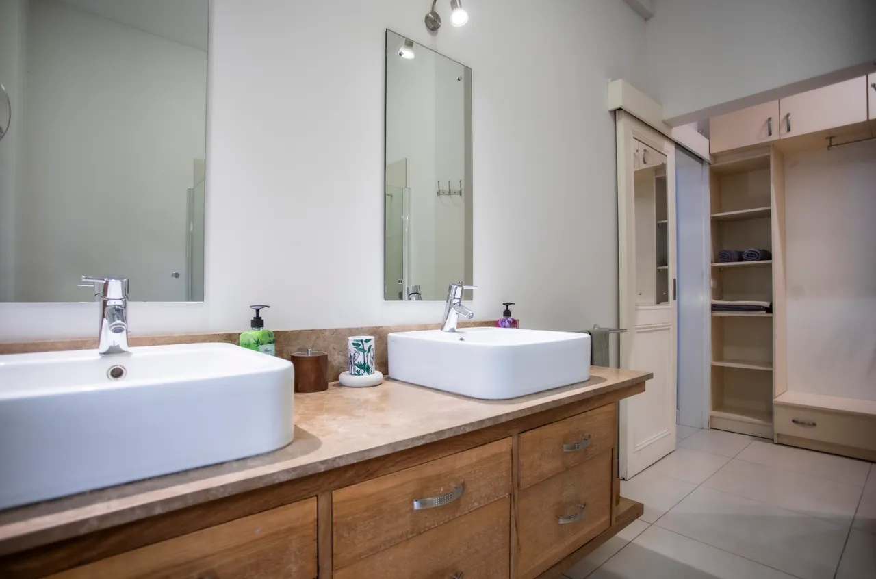 Espace salle de bain avec doubles lavabos sur un plan en bois.  
Miroirs éclairés au-dessus des vasques pour un éclairage optimal.  
Rangement intégré avec étagères pour les accessoires de toilette.  
Douche à l'italienne en verre, offrant une utilisation fonctionnelle.  
Fenêtre apportant lumière naturelle et ventilation à l'espace.