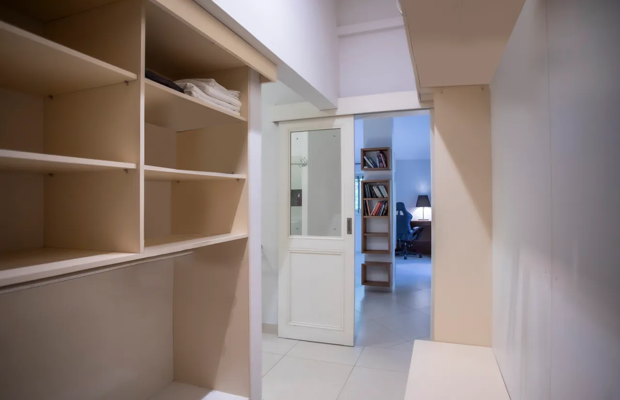 Espace de rangement avec étagères intégrées et coin bureau à l'arrière.  
Sol carrelé clair contribuant à une ambiance spacieuse et fonctionnelle.  
Porte vitrée permettant l'accès à la pièce adjacente.  
Ambiance lumineuse favorisée par les murs aux teintes neutres.  
Rangement optimisé avec des étagères ouvertes et un