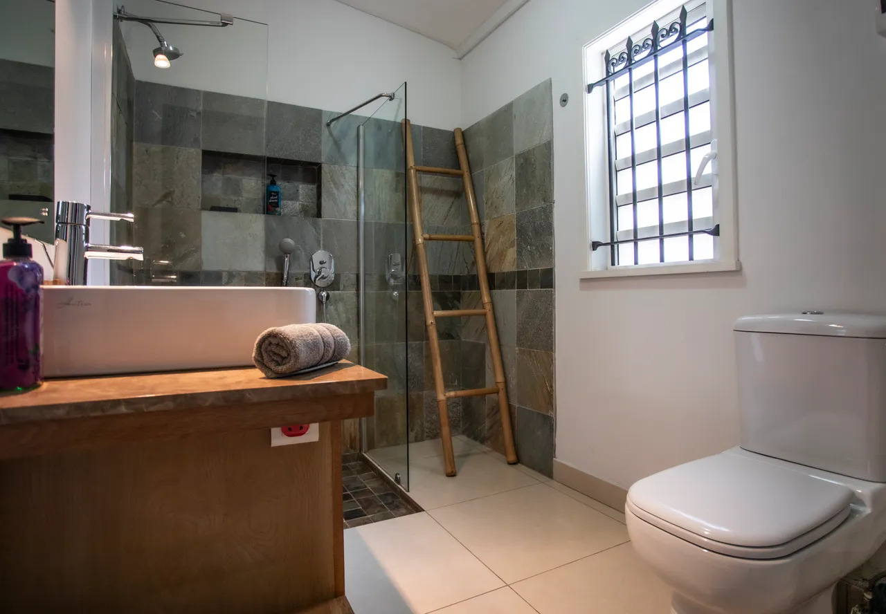 Salle de bain avec douche à l'italienne et lavabo en bois.  
Miroir éclairé au-dessus du lavabo pour un éclairage fonctionnel.  
Étagères en bois offrent un espace de rangement pratique.  
Fenêtre avec grille permet ventilation et lumière naturelle.  
Sol carrelé clair contribue à une atmosphère spacieuse.