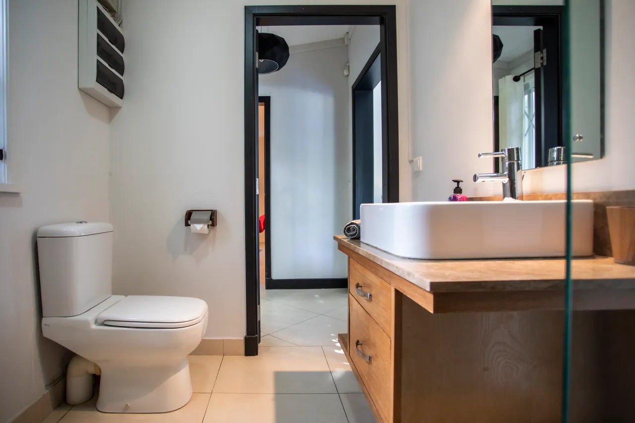 Salle de bain équipée d'un lavabo moderne sur un meuble en bois.  
Miroir au-dessus de la vasque avec éclairage intégré pour une meilleure visibilité.  
Fenêtre offrant lumière naturelle tout en permettant l'aération.  
Douche à l'italienne en verre pour un accès pratique.  
Sol carrelé clair favorise une ambiance spacieuse et lumineuse.