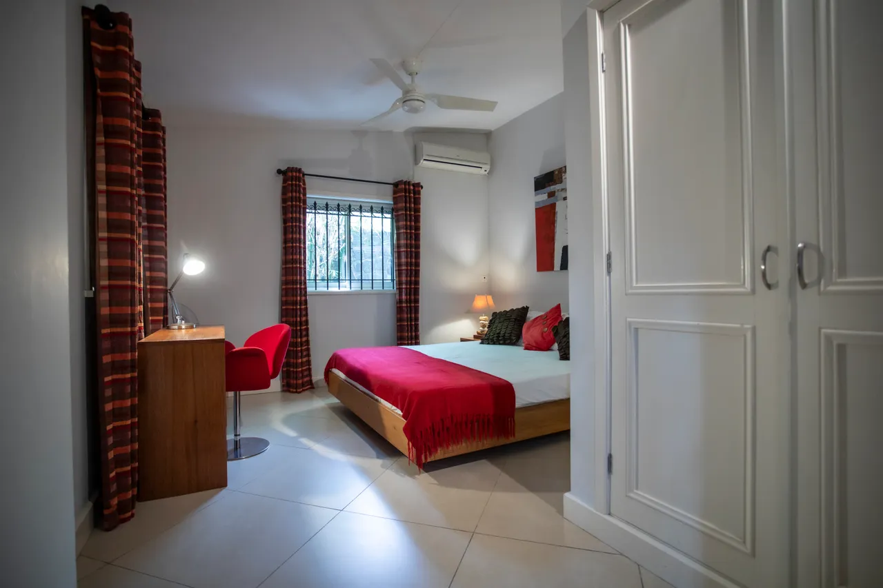 Chambre lumineuse avec lit en bois et grands rideaux à motifs.  
Coin bureau avec chaise rouge et lampe de travail pratique.  
Fenêtre offrant une vue sur l'extérieur et apportant de la lumière.  
Rangement intégré avec placard pour un espace fonctionnel et ordonné.  
Sol carrelé clair contribue à une ambiance aérée et spacieuse.