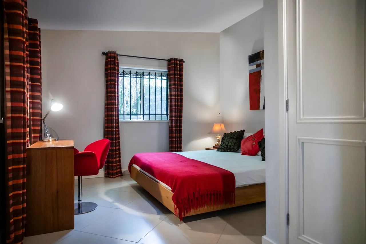 Espace avec lit double et lumière douce des lampes de chevet.  
Fenêtre avec barreaux offre une vue sur l'extérieur.  
Coin bureau équipé d'une chaise rouge et d'une lampe de travail.  
Rideaux à motifs ajoutent une touche de couleur et d'intimité.  
Sol carrelé clair renforce une ambiance spacieuse et lumineuse.