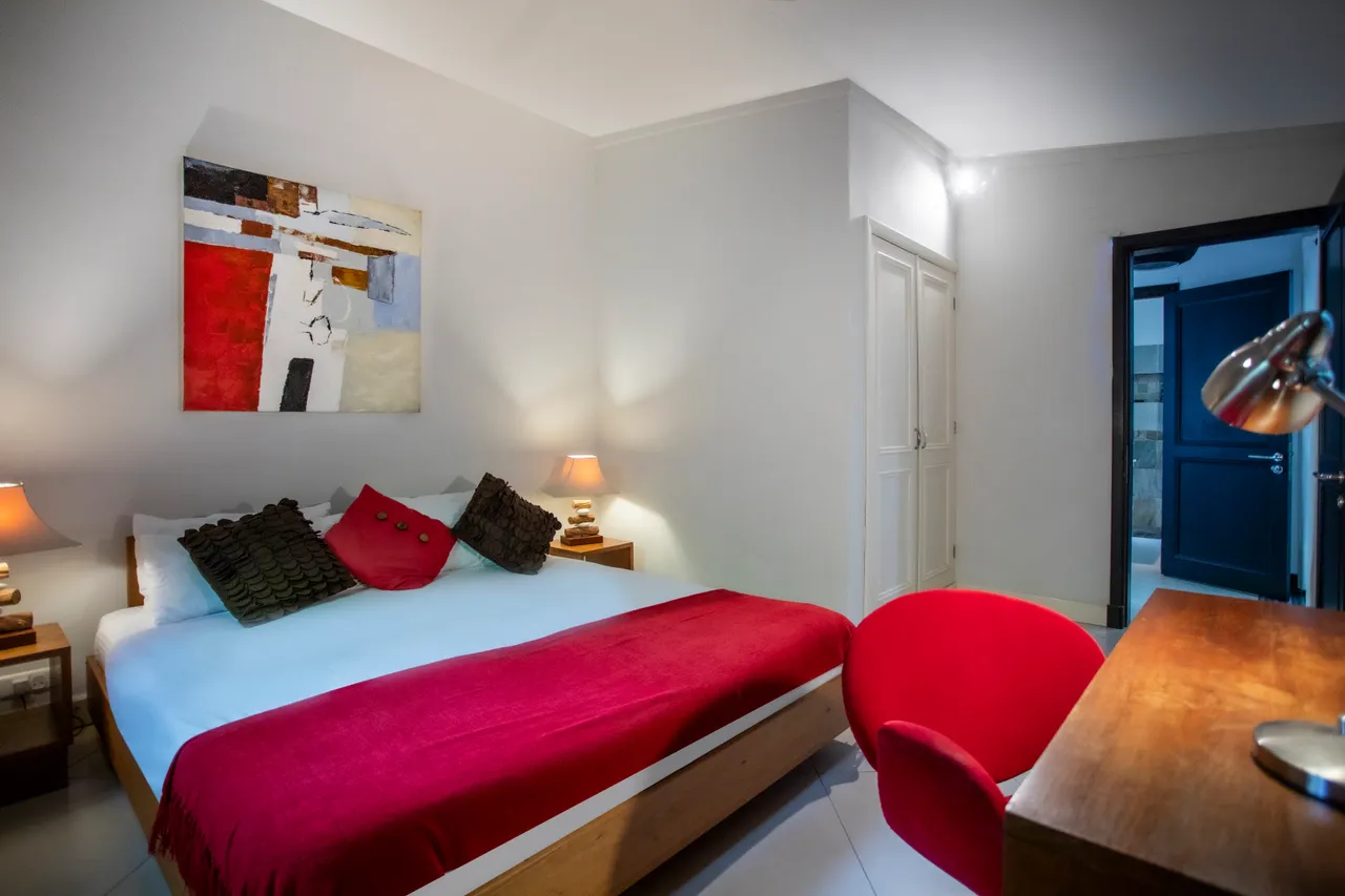 Tons neutres créent une atmosphère sereine dans la chambre.  
Lit double agrémenté de coussins colorés pour un confort simple.  
Coin bureau en bois avec lampe de travail pour un espace fonctionnel.  
Fenêtre laisse entrer la lumière naturelle tout en offrant une vue.  
Sol carrelé clair contribue à l'impression d'espace dans la pièce.