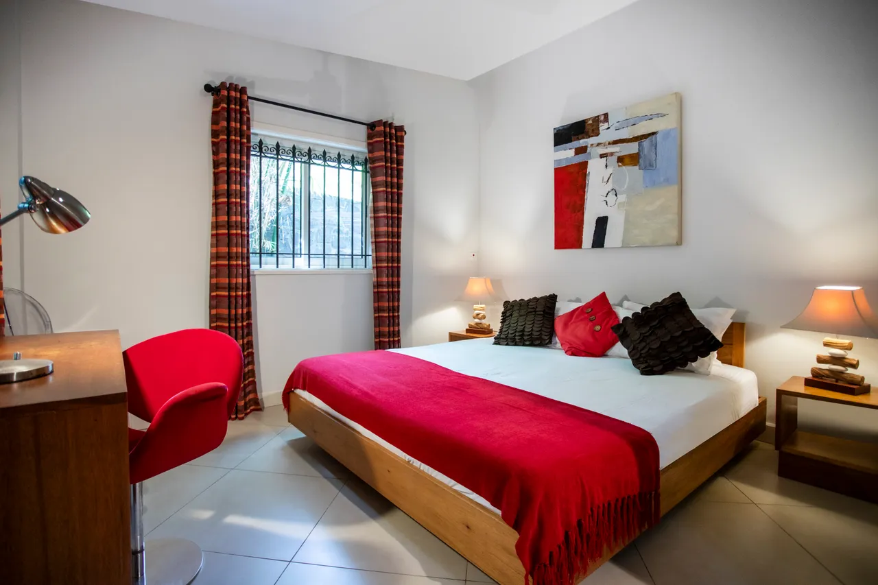Chambre équipée d'un lit double avec linge de lit en coton.  
Mobilier en bois avec lampes de chevet pour un éclairage fonctionnel.  
Fenêtre avec barreaux offrant une vue sur l'extérieur.  
Sol carrelé clair renforçant l'impression d'espace et de luminosité.  
Rideaux à motifs ajoutent une touche de couleur à la décoration.