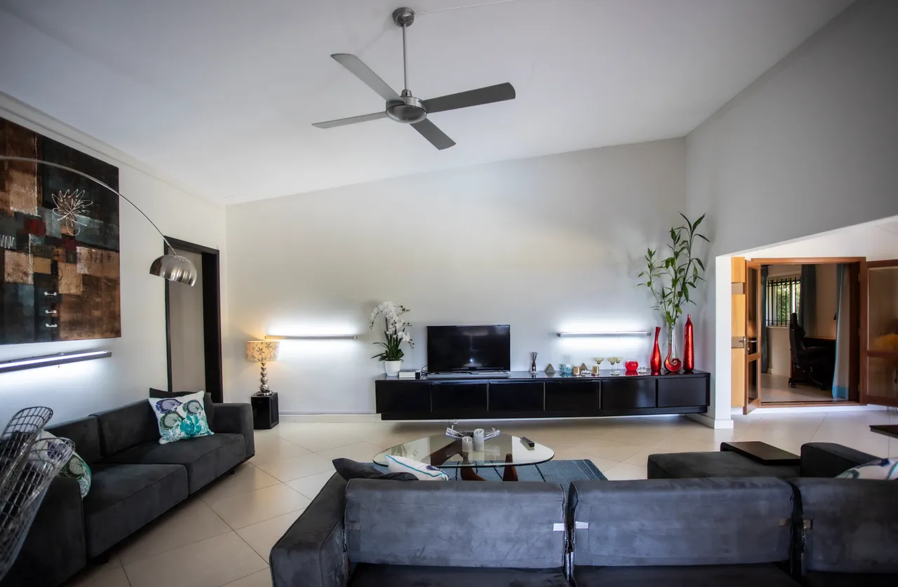 Canapé en tissu gris et table en verre créent un espace convivial.  
Éclairage indirect avec appliques murales accentue l'ambiance chaleureuse.  
Grand téléviseur sur meuble noir, optimisant l'espace de divertissement.  
Ventilateur au plafond assure une circulation d'air agréable dans la pièce.  
Sol carrelé clair et murs neutres renforcent la luminosité