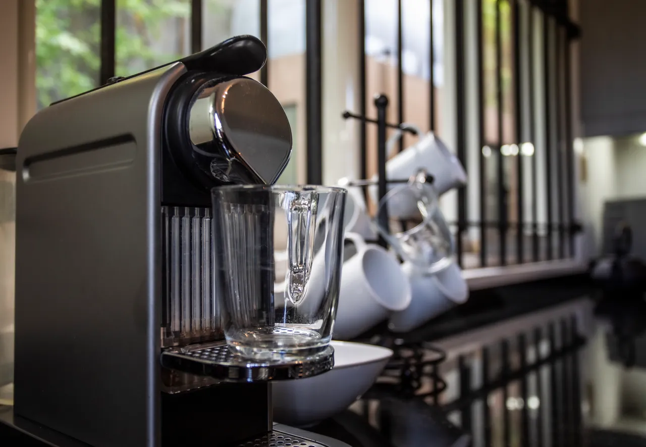 Espace cuisine équipé d'une machine à café moderne et de vaisselle.  
Plans de travail en granit facilitent la préparation des repas.  
Fenêtre avec grille permet une ventilation et une lumière naturelle.  
Rangement optimal avec placards et étagères pour la vaisselle.  
Sol carrelé clair contribue à une atmosphère propre et spacieuse.