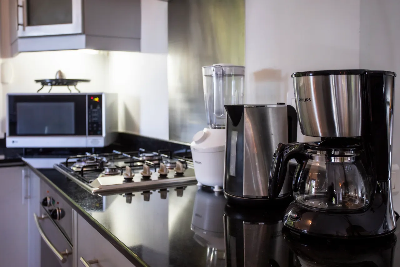 Espace cuisine avec appareils modernes, dont un mixeur et un four à micro-ondes.  
Plans de travail en granit offrant une surface pratique pour cuisiner.  
Rangement avec placards et étagères pour organiser la vaisselle.  
Fenêtre apportant lumière naturelle et ventilation à l'espace.  
Sol carrelé clair favorise une ambiance propre et accueillante.