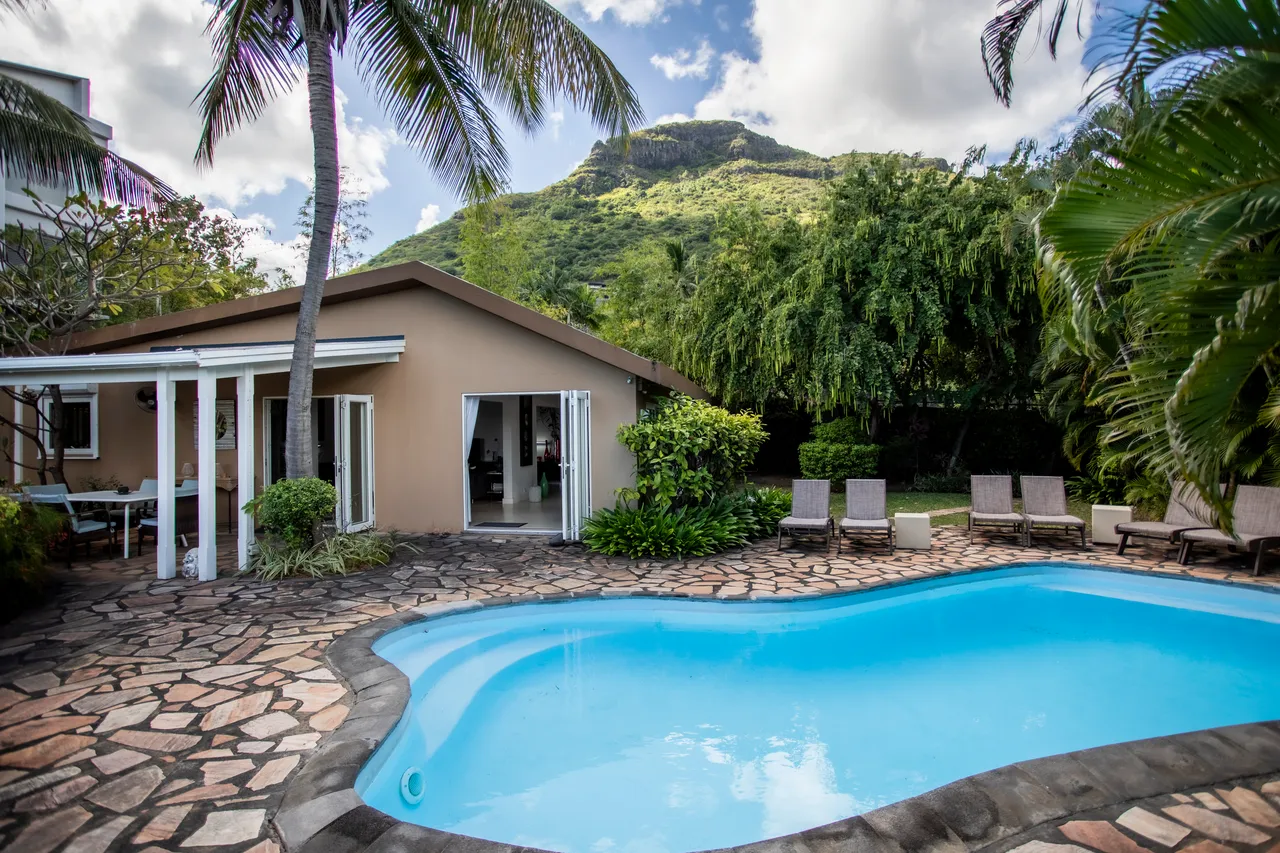 Espace extérieur avec piscine privée entourée de transats.  
Terrasse dallée offrant un accès direct à la villa.  
Végétation tropicale ajoute une touche naturelle à l'environnement.  
Vue dégagée sur la montagne depuis le jardin.  
Palmier en arrière-plan contribue à l'ambiance tropicale.