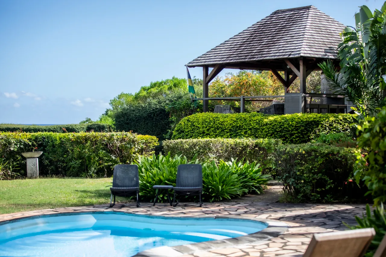 Espace extérieur avec piscine et transats, idéal pour se détendre.  
Pergola en bois offre un coin ombragé pour se ressourcer.  
Végétation dense encadre le jardin, apportant intimité et verdure.  
Terrasse dallée facilite l'accès à la piscine et à la villa.  
Vue dégagée sur le jardin et le paysage environnant.