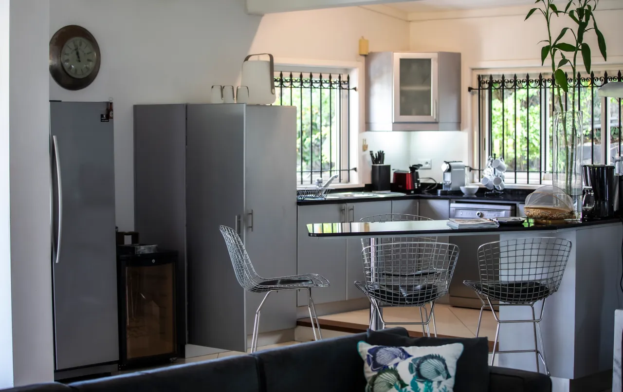 Cuisine moderne avec bar en granit et chaises design.  
Équipement électroménager complet pour une utilisation pratique.  
Fenêtres avec grilles permettant une bonne aération et lumière.   
Rangements optimisés avec placards intégrés pour les ustensiles.  
Sol carrelé clair facilitant l'entretien et apportant de la luminosité.