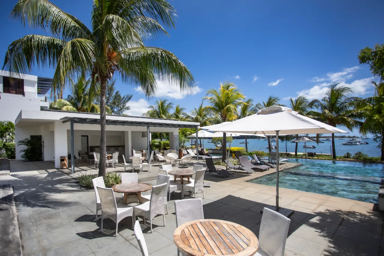 Le carrelage gris entoure la piscine, bordée de tables, de chaises et d’un parasol.