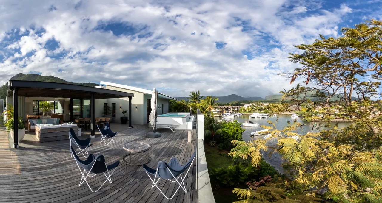 Terrasse en bois avec vue sur le port et les montagnes environnantes.  
Coin détente avec chaises longues et parasol.  
Espace barbecue intégré pour des repas en plein air.  
Végétation variée apportant une touche naturelle à l'environnement.  
Canapé d'extérieur offrant un confort pour se relaxer au soleil.  
Grande baie vitrée connectant l'intérieur à l