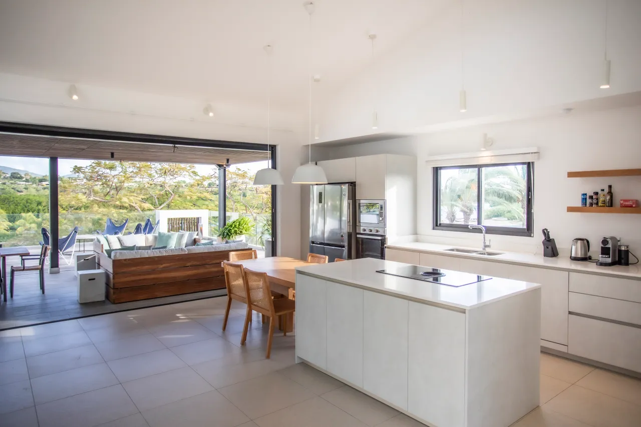 Espace de cuisine moderne avec îlot central et équipements intégrés.  
Salle à manger adjacente offrant une vue sur l'extérieur.  
Éclairage suspendu discret ajoutant une touche contemporaine.  
Mobilier en bois crée un contraste chaleureux avec le design moderne.  
Accès direct à la terrasse pour des repas en plein air.