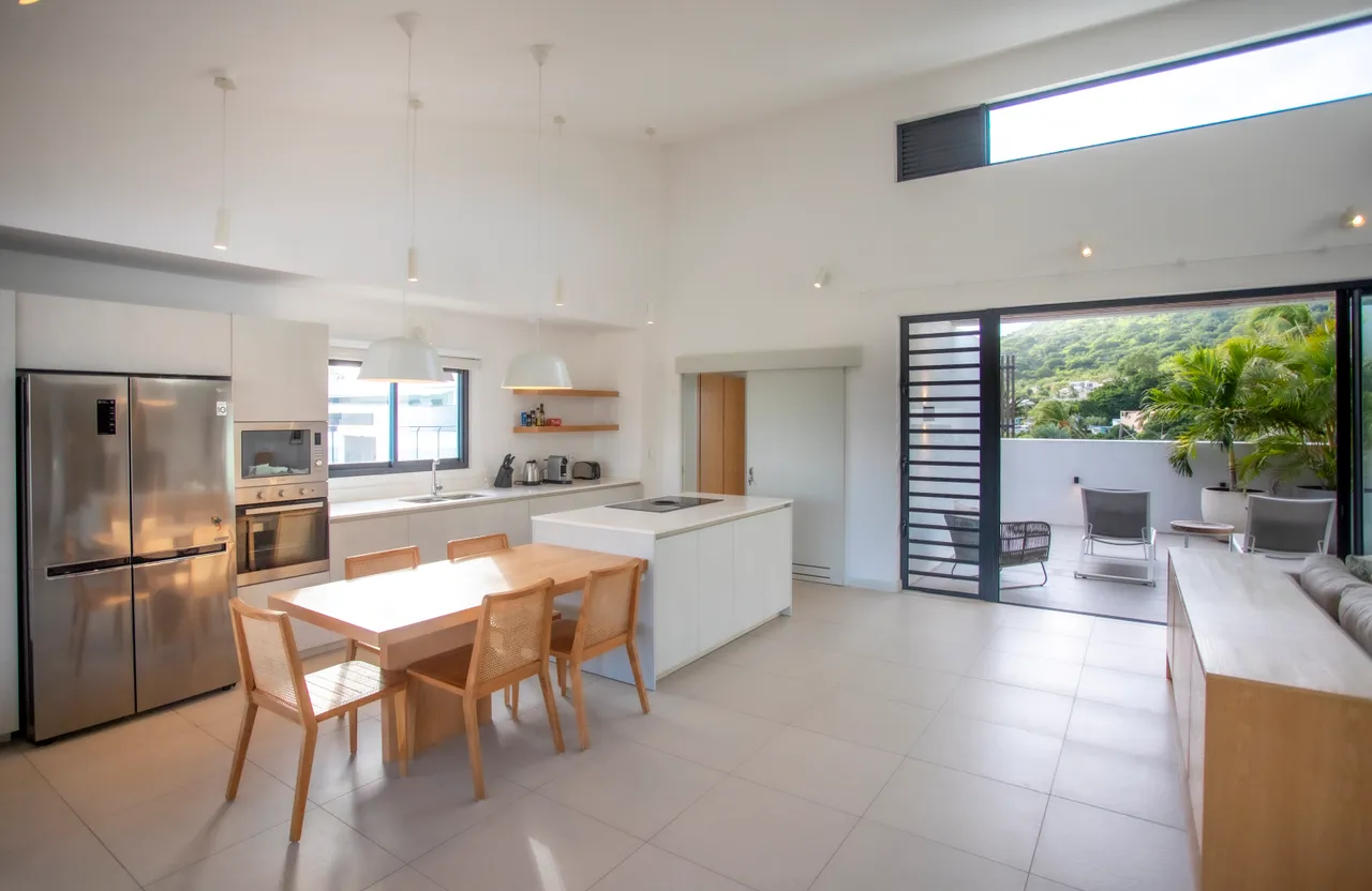 Cuisine ouverte avec îlot central et équipements modernes.  
Mobilier en bois clair et chaises assorties créent une harmonie.  
Lumière naturelle abondante grâce aux grandes fenêtres.  
Espace de repas adjacent avec vue sur l'extérieur.  
Accès direct à la terrasse pour des repas en plein air.