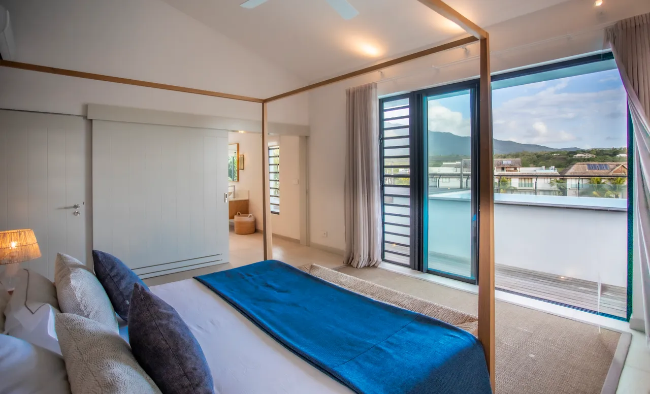 Tons neutres et texture douce du linge de lit créent une atmosphère calme. Grand balcon offrant une vue sur l'extérieur et la nature. Lit à baldaquin ajoutant une touche de sophistication à la pièce. Éclairage naturel abondant grâce aux grandes fenêtres. Mobilier minimaliste favorisant un espace aéré et agréable.