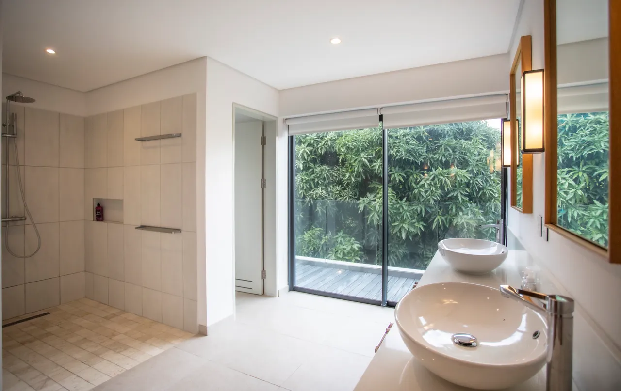 Salle de bain avec carrelage clair et grande douche à l'italienne.  
Double vasque contemporaine en matériaux épurés.  
Fenêtre donnant sur la végétation extérieure, apportant lumière naturelle.  
Éclairage intégré créant une ambiance chaleureuse et moderne.  
Rangement discret sous le plan de travail pour un espace dégagé.
