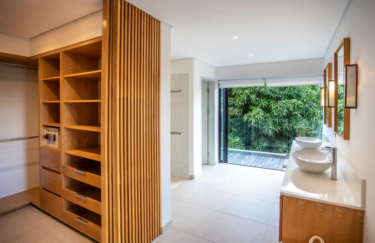 Salle de bain avec double vasque et miroir élégant. Rangement intégré en bois optimisant l'espace. Fenêtre ouvrante offrant une vue sur la végétation. Éclairage moderne créant une ambiance accueillante. Douche à l'italienne avec carrelage clair pour une atmosphère épurée.