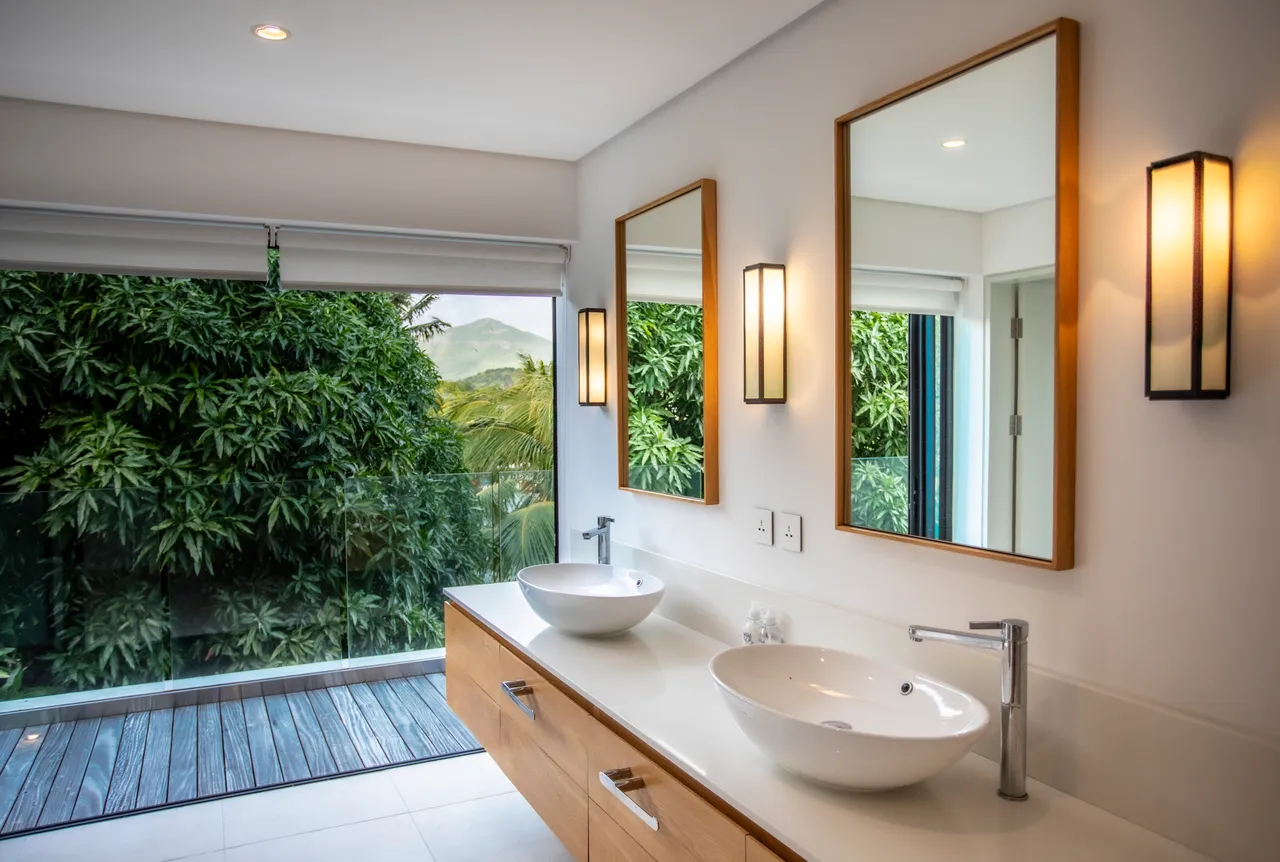 Salle de bain avec double vasque et éclairage moderne. Rangement intégré en bois pour un espace fonctionnel. Grandes fenêtres offrant une vue sur la végétation environnante. Miroirs ajoutent de la profondeur à l'espace. Douche à l'italienne avec carrelage clair pour un design épuré.