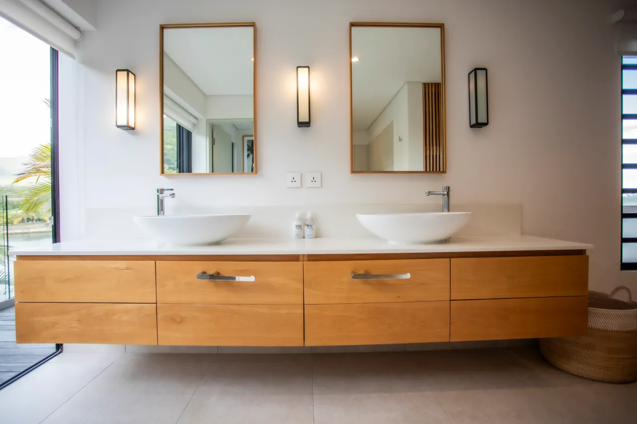 Teintes claires et matériaux naturels dominent la salle de bain. Double vasque au design épuré avec miroir encadré. Rangement en bois optimisant l'espace tout en restant fonctionnel. Fenêtre apportant lumière naturelle et vue sur l'extérieur. Douche à l'italienne avec carrelage clair pour une ambiance contemporaine.