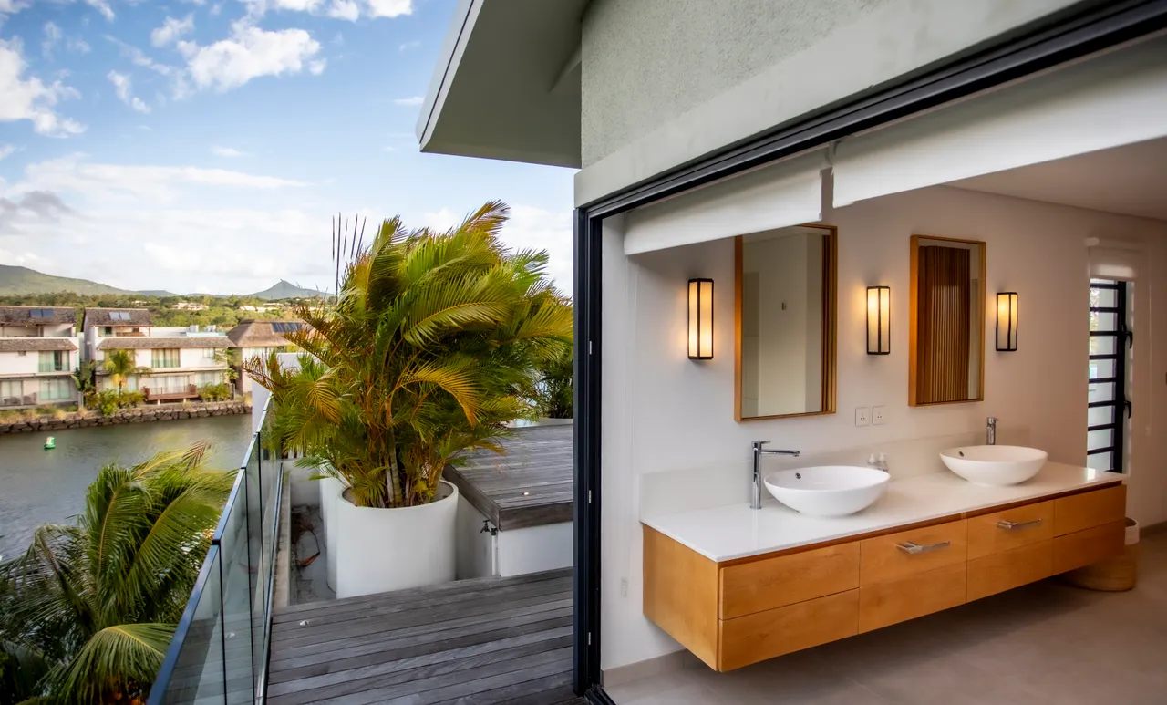 Salle de bain avec double vasque et miroir en bois. Rangement en bois discret optimisant l'espace. Fenêtre ouvrante offrant une vue sur la terrasse. Éclairage indirect créant une atmosphère douce et moderne. Douche à l'italienne avec carrelage clair.