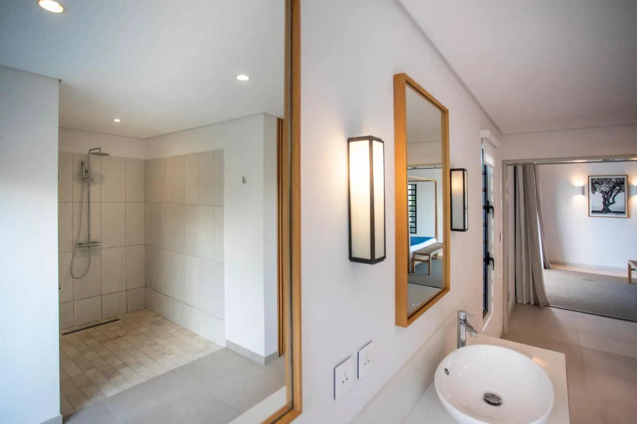 Salle de bain avec douche à l'italienne et carrelage clair. Miroir encadré ajoutant une touche moderne. Éclairage stratégique offrant une ambiance agréable. Rangement intégré en bois optimisant l'espace disponible. Grande fenêtre apportant lumière naturelle et vue extérieure.