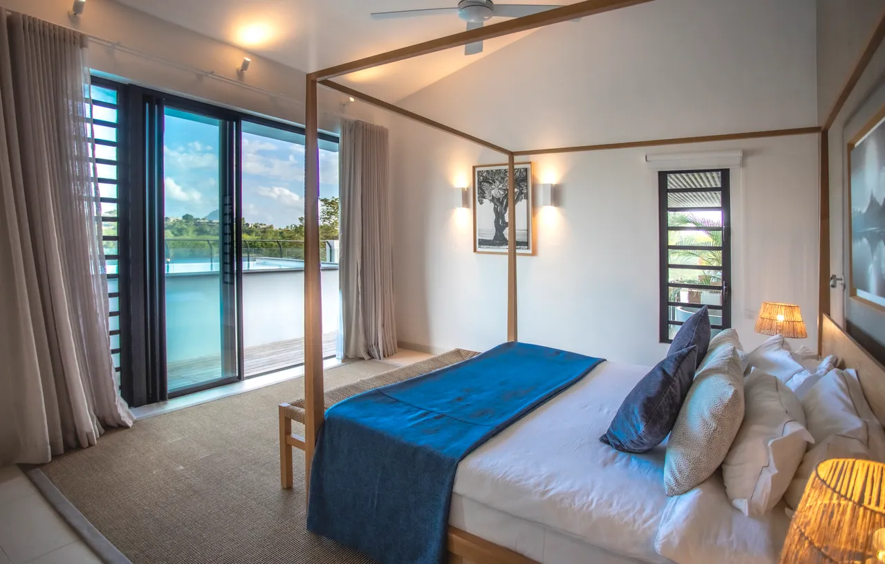 Chambre lumineuse avec accès direct à un balcon.  
Lit à baldaquin et décoration minimaliste.  
Vues dégagées depuis les grandes fenêtres.  
Tapis en jute apportant une texture naturelle à l'espace.  
Linge de lit aux teintes douces pour une ambiance sereine.
