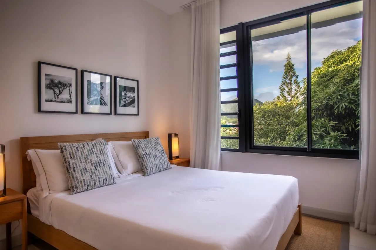 Chambre avec vue sur la végétation environnante à travers une grande fenêtre. 

Lit en bois avec éclairage d'appoint créant une ambiance douce.

Décoration murale en noir et blanc apportant une touche artistique. 

Tons clairs des murs favorisant une atmosphère apaisante. 

Rangement intégré optimisant l'espace disponible dans la pièce.