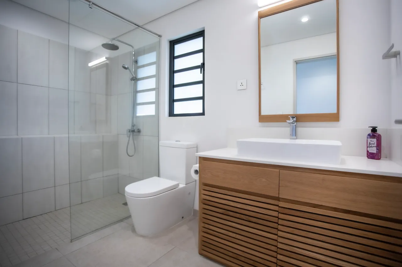 Salle de bain équipée d'une douche à l'italienne et d'un meuble en bois. 

Miroir minimaliste et éclairage intégré renforçant la fonctionnalité. 

Fenêtre étroite apportant lumière naturelle sans compromettre l'intimité. 

Carrelage clair offrant un aspect moderne et lumineux.